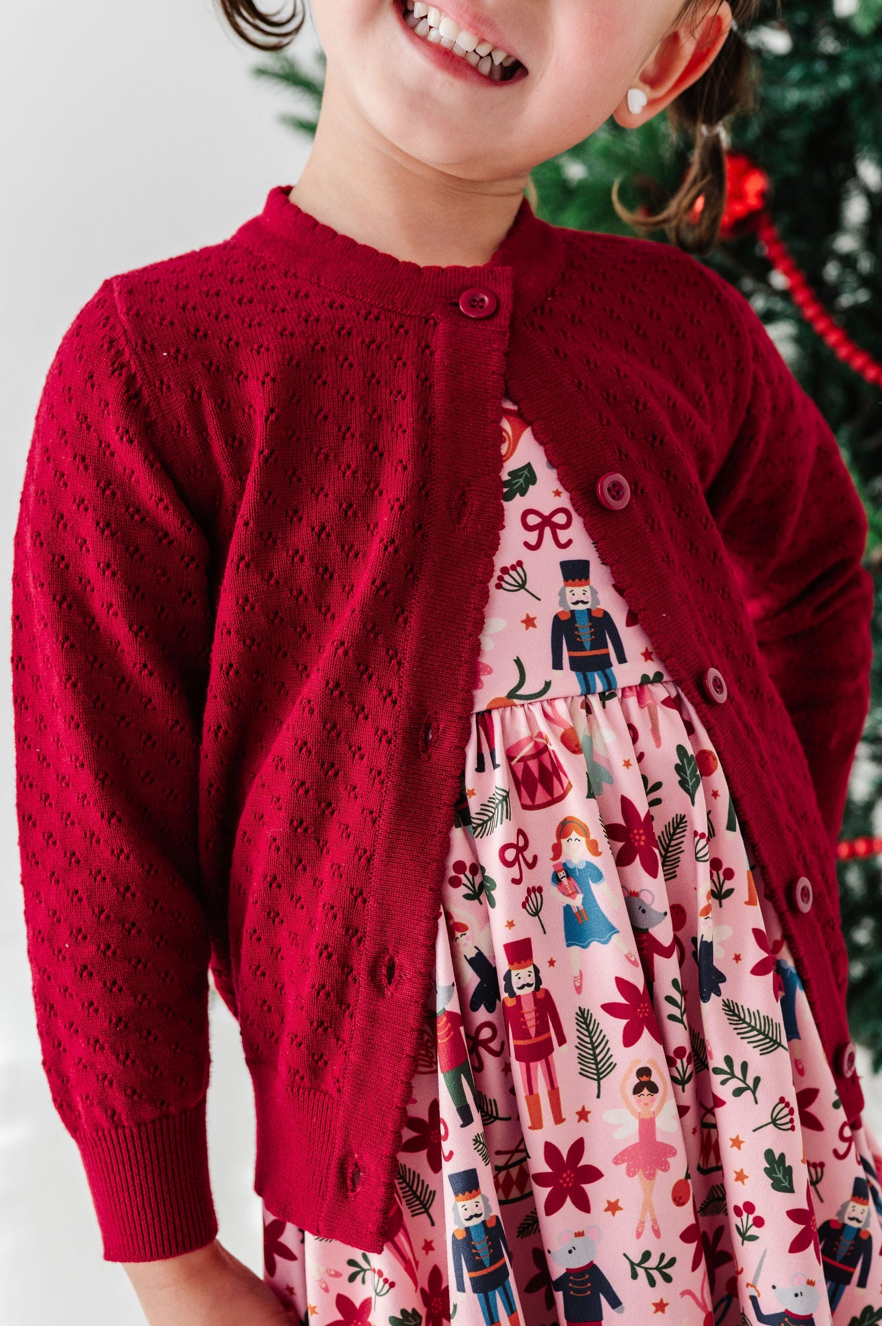 A young child wearing a festive holiday outfit, featuring a deep red knit cardigan layered over a pink dress with a colorful nutcracker and holiday pattern, set against a softly blurred Christmas tree background.