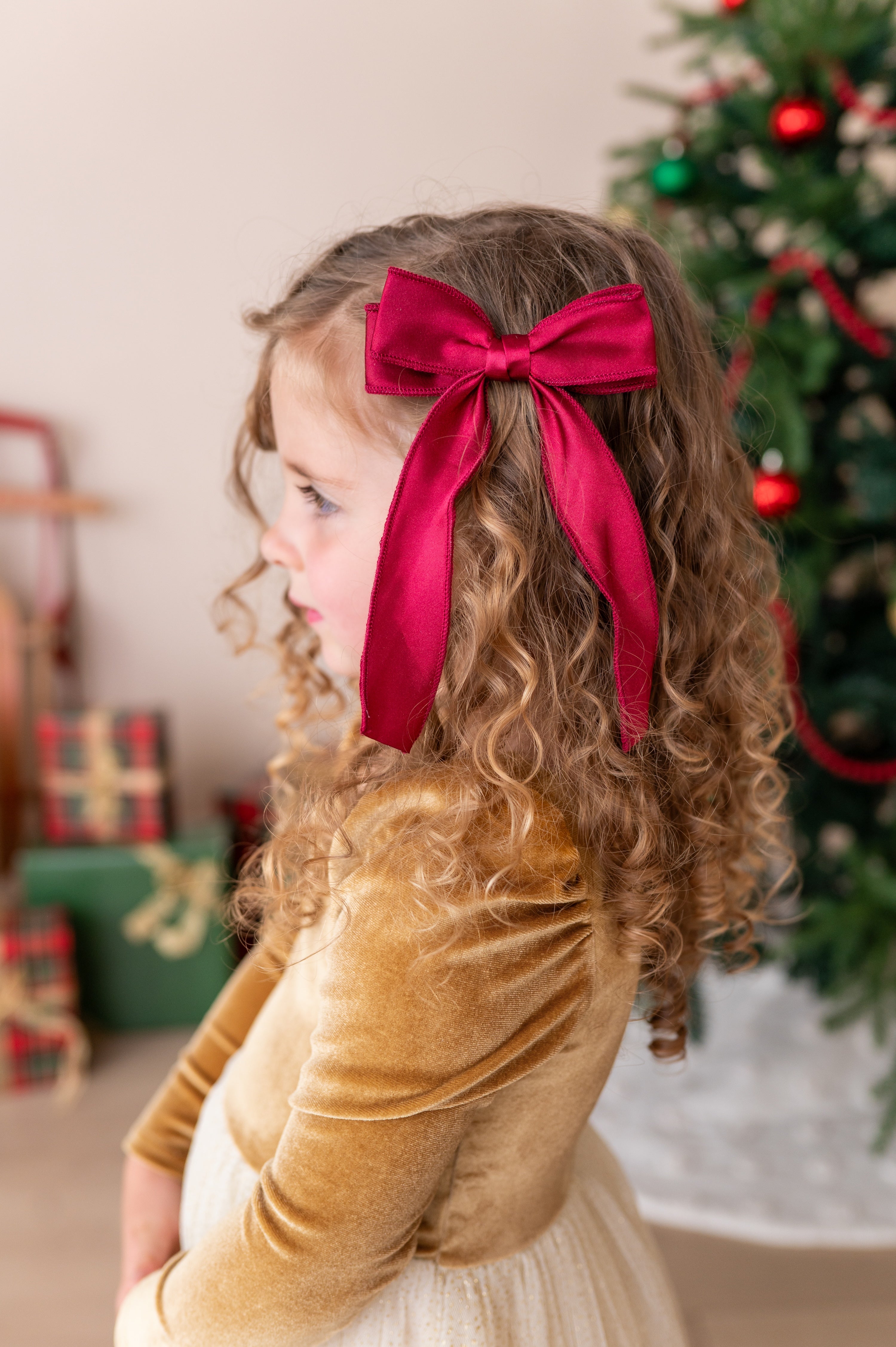 A young girl with curly brown hair wearing a large, bright red satin bow headband, dressed in a gold velvet top and cream skirt, standing in front of a decorated Christmas tree.