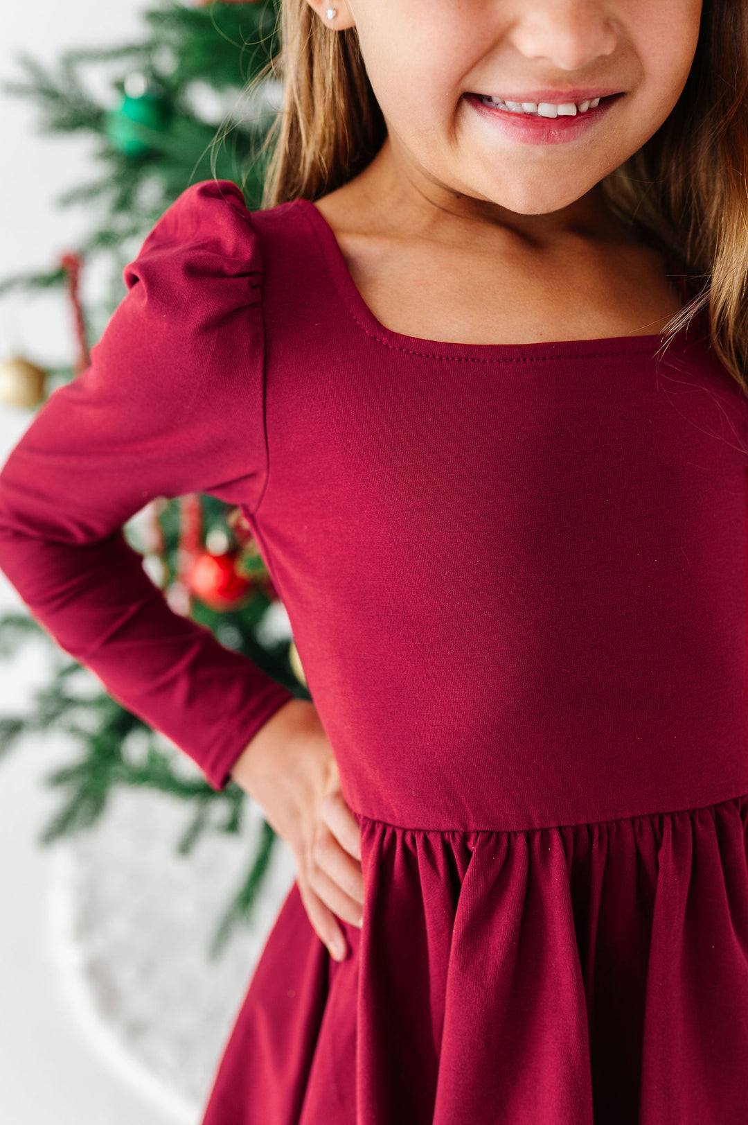 A close-up of a young girl wearing a long-sleeve burgundy dress with a square neckline and puffed shoulders, standing in front of a blurred Christmas tree.
