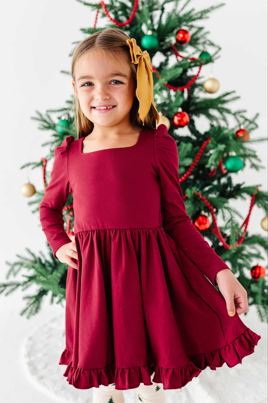 A young girl wearing a long-sleeve burgundy dress with a square neckline and ruffled hem, standing in front of a decorated Christmas tree.