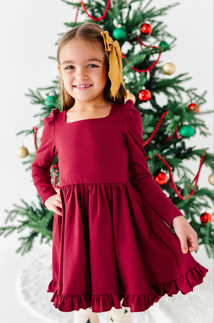 A young girl wearing a long-sleeve burgundy dress with a square neckline and ruffled hem, standing in front of a decorated Christmas tree.