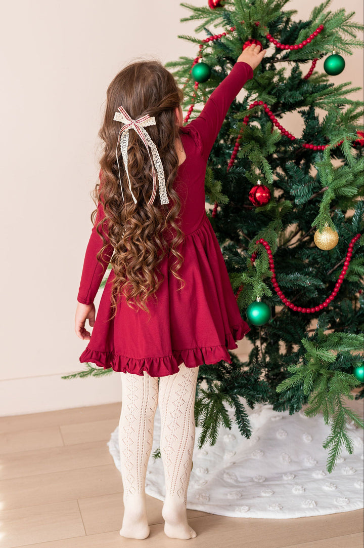 A young girl with long wavy brown hair is seen from behind, wearing a long-sleeve burgundy dress, cream cable-knit tights, and a white hair bow, reaching to decorate a Christmas tree with red and green ornaments.