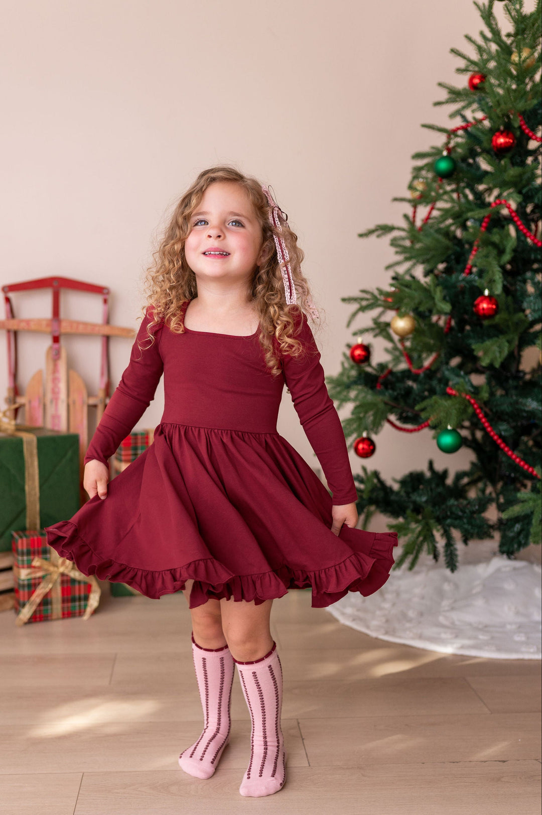 A young girl with curly blonde hair wearing a long-sleeve burgundy dress and pink striped socks, standing in a festive room next to a decorated Christmas tree and wrapped gifts.