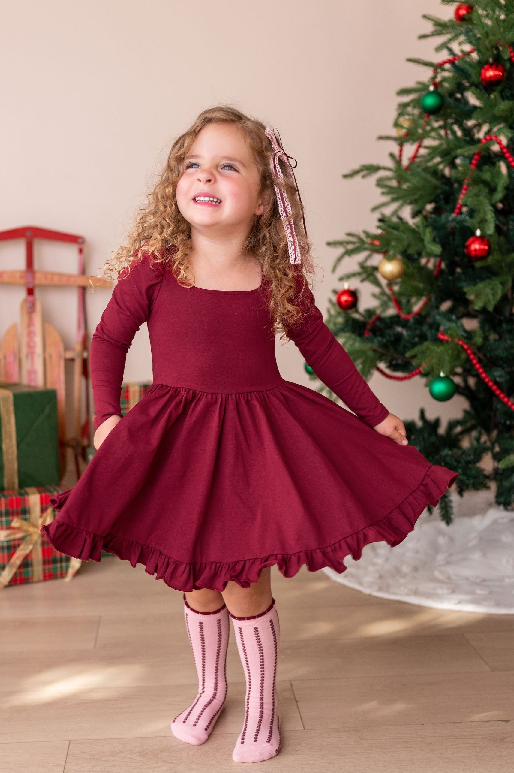 A young girl with curly blonde hair wearing a long-sleeve burgundy dress and pink striped socks, posing in front of a decorated Christmas tree and wrapped gifts.