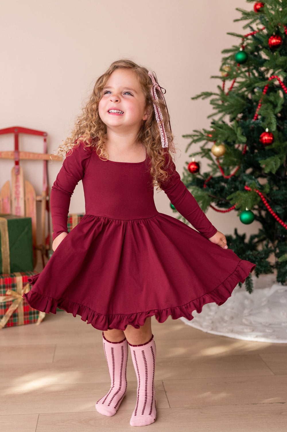 A young girl with curly blonde hair wearing a long-sleeve burgundy dress and pink striped socks, posing in front of a decorated Christmas tree and wrapped gifts.