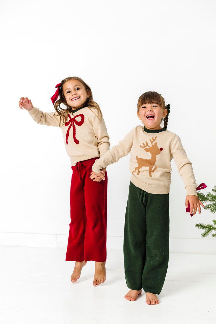 children jumping in cream Christmas sweaters with red bow and brown reindeer designs and coordinating cotton knit pants in red and green