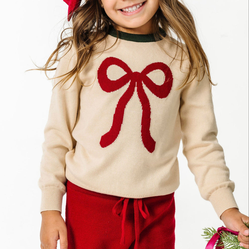 little girl in cream sweater with red bow design paired with matching red sweater pants and satin bow for Christmas