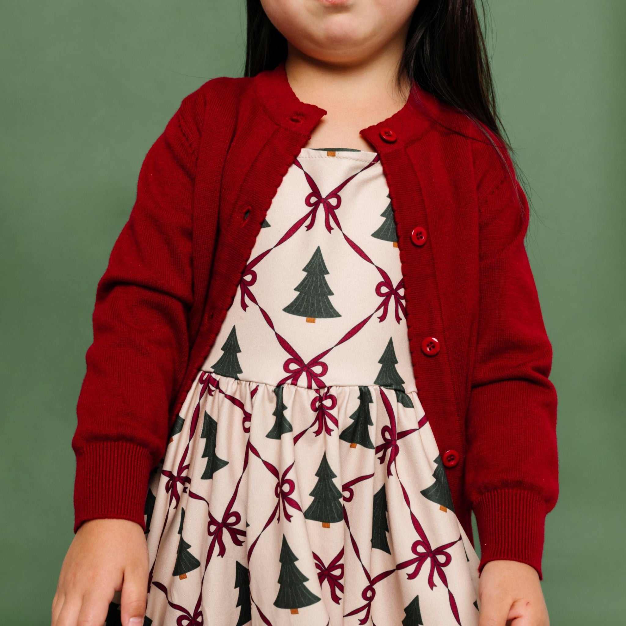 little girl in Christmas tree and bow print twirl dress with deep red cotton knit cardigan