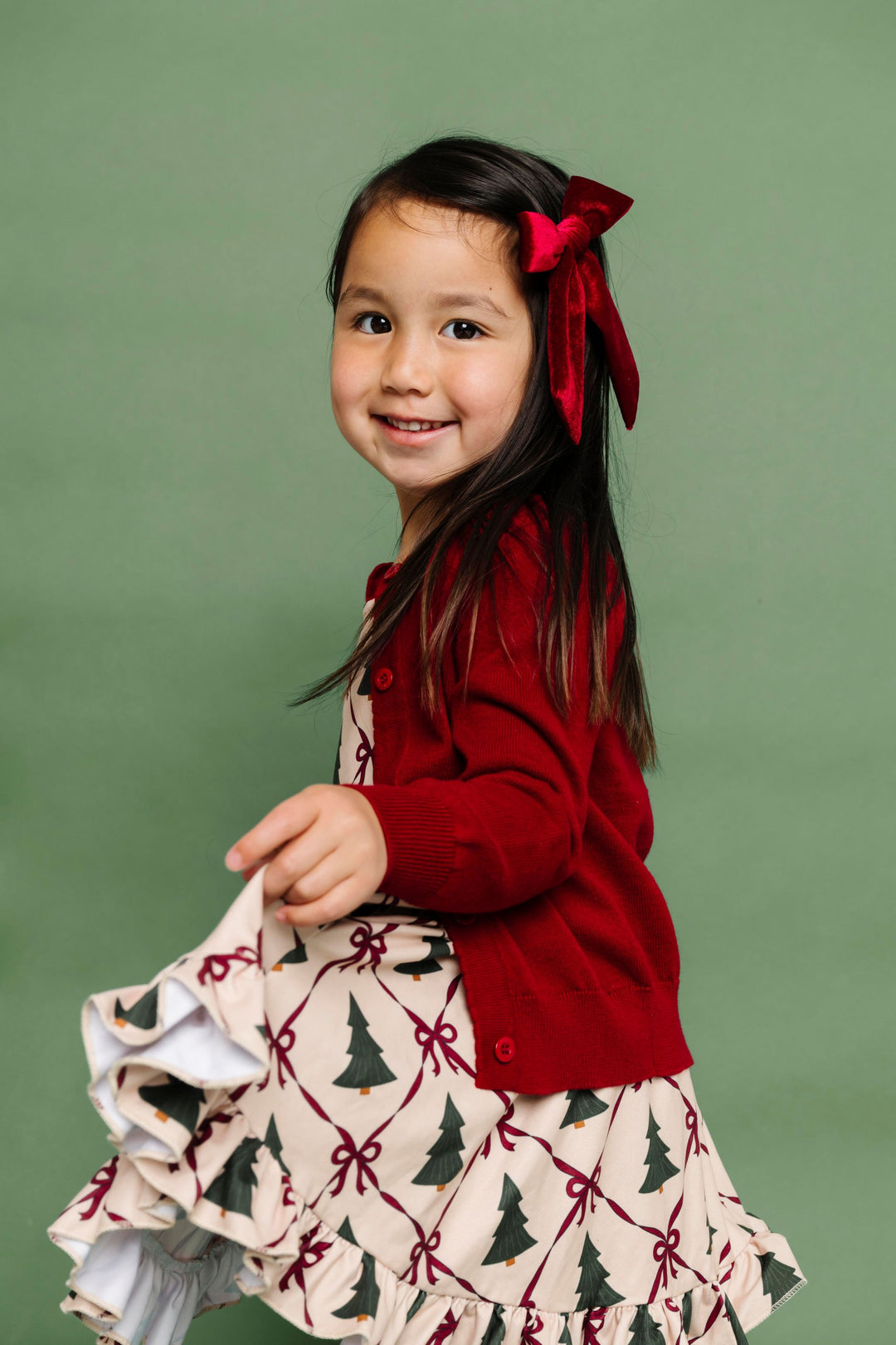 little girl in cream dress with Christmas tree and bow print paired with matching crimson button front cardigan and velvet bow