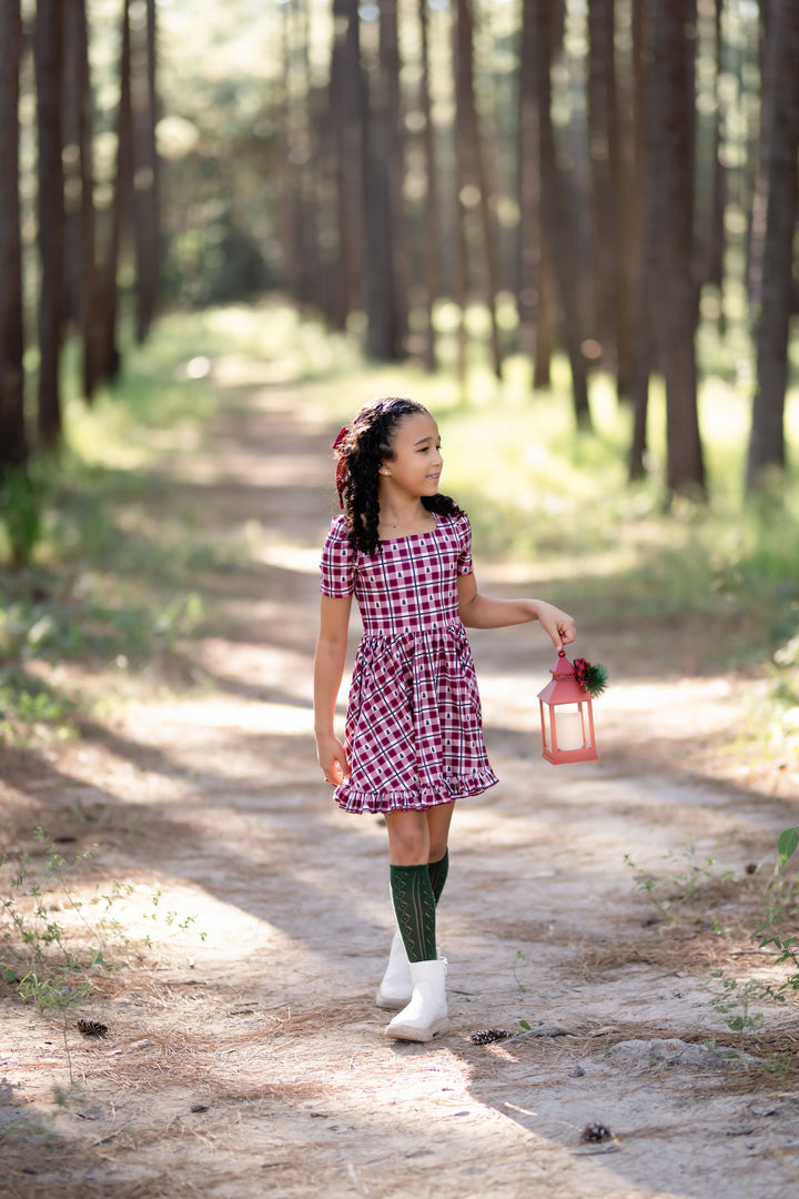 little girl in red gingham Christmas tree dress with matching dark green open knit knee highs in the forest