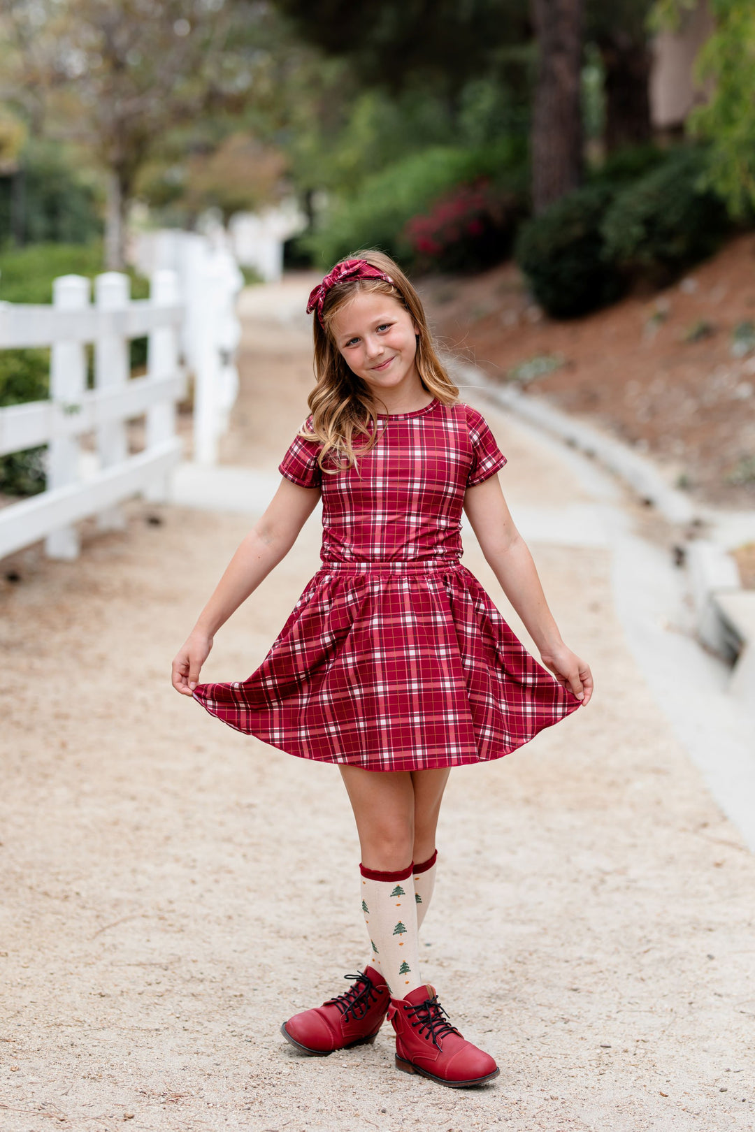 young girl in matching headband, top and skirt in crimson plaid paired with Christmas tree print knee high socks and red boots