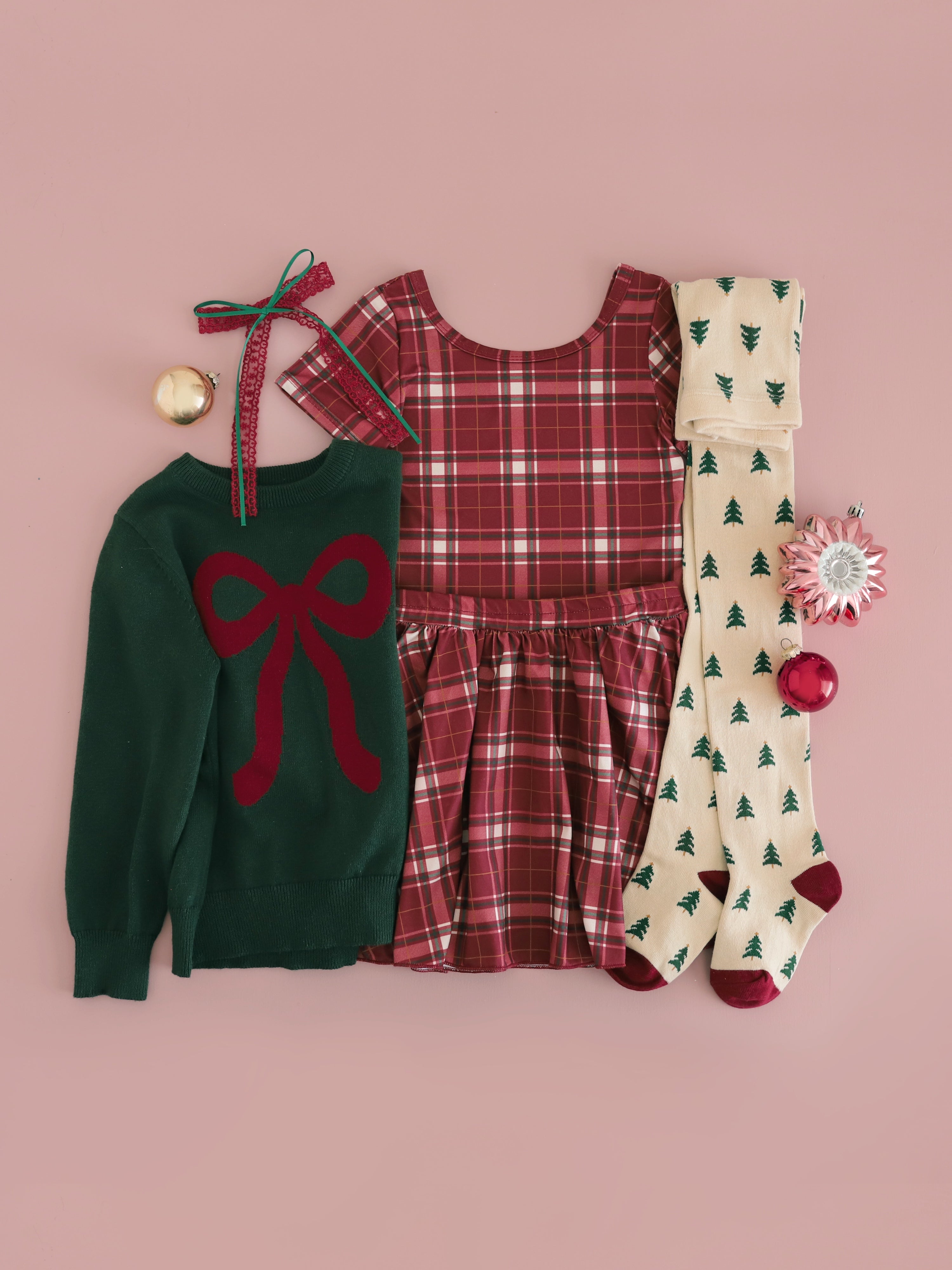 flat lay of girls' crimson plaid top and skirt set paired with forest green pullover with crimson bow design and matching cream tights with Christmas trees 