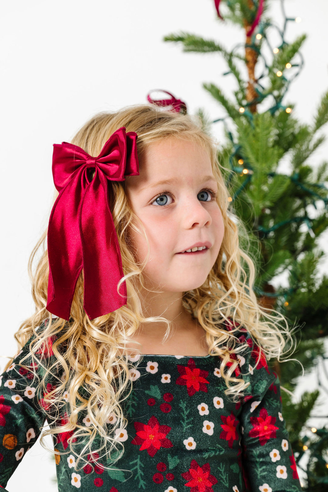 girl by Chrismas tree in crimson satin double hair bow on clip paired with poinsettia print velvet dress