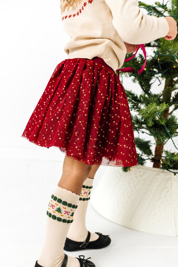 little girl decorating Christmas tree in crimson tulle skirt with gold sparkly dots paired with cream fair isle print knee high socks