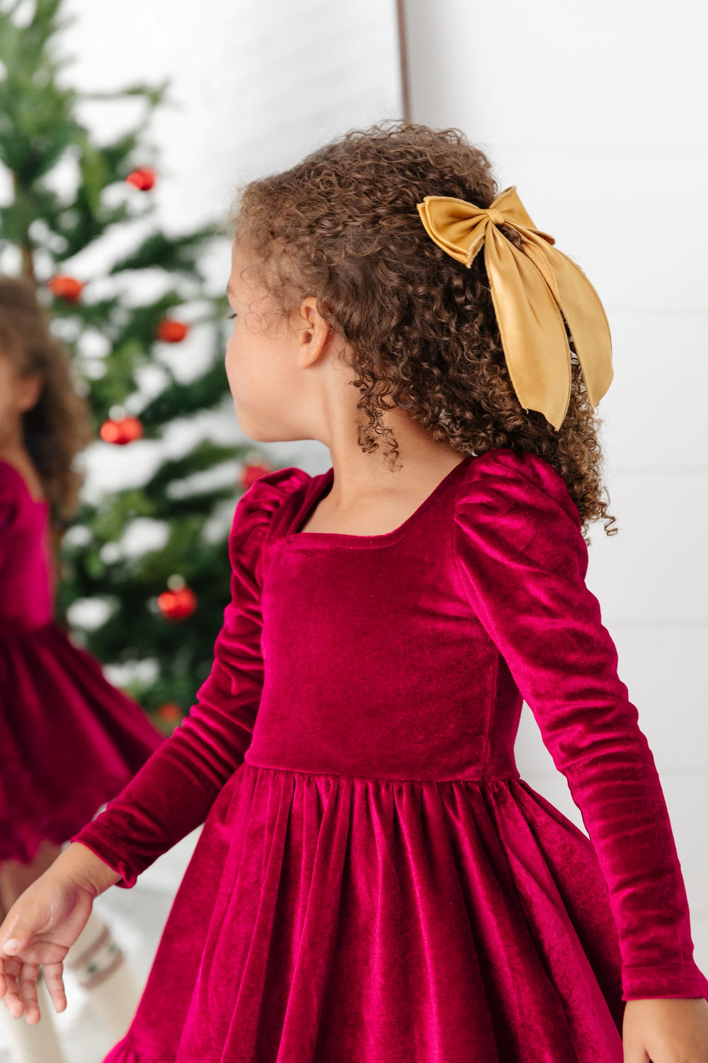 young girl looking in mirror in deep red velvet long sleeve twirl dress with oversized gold satin hair bow on clip for Christmas