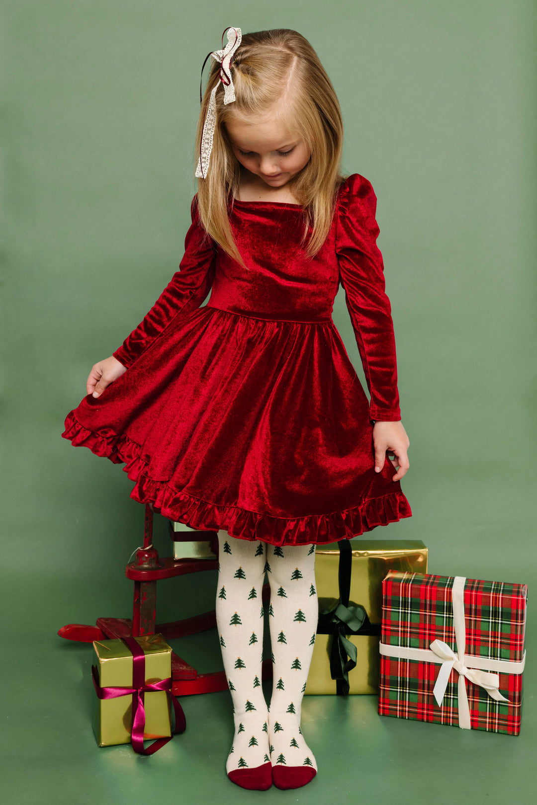 little girl in front of Christmas gifts wearing crimson velvet twirl dress with long sleeves paired with cream lace bow and cream tights with tree pattern