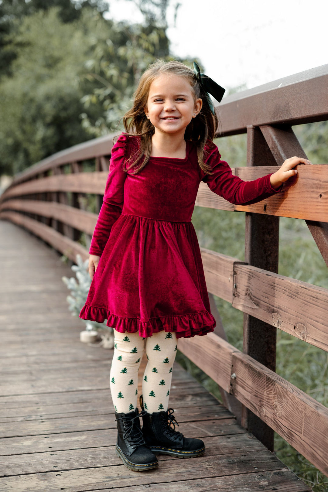 little girl on woodland bridge in cranberry red velvet long sleeve Christmas dress and Christmas tree tights