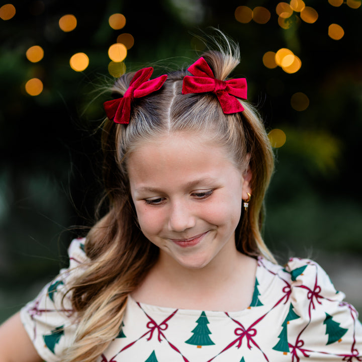 girl in front of Christmas lights with deep red velvet pigtail bows on clips paired with matching cream twirl dress with Christmas tree and crimson ribbon print