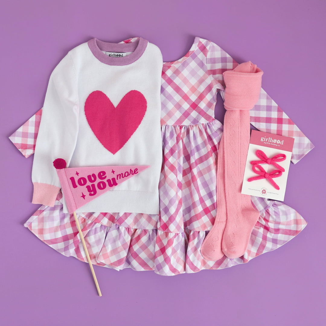 girl's Valentine's Day outfit inspiration in shades of pink and purple with matching gingham dress, heart sweater, light pink open knit tights and pink yarn pigtail bows