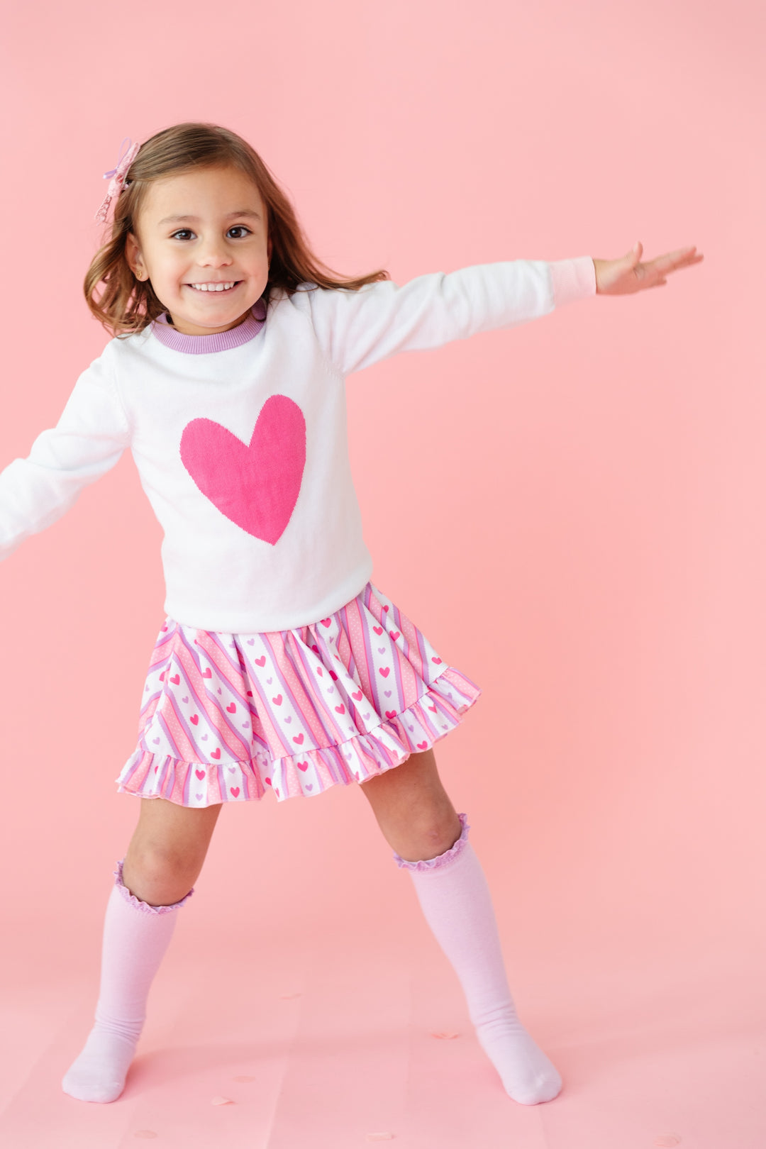 little girl in white cotton knit pullover sweater with pink heart paired with matching pink wallpaper inspired twirl dress and lavender knee highs