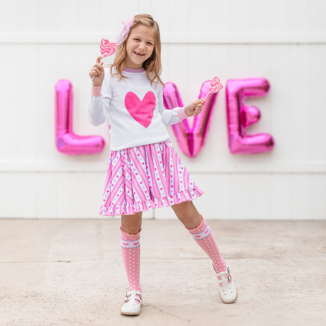 little girl holding heart lollipops in matching heart sweater, wallpaper inspired twirl dress and pink knee high socks with polka dots and hearts