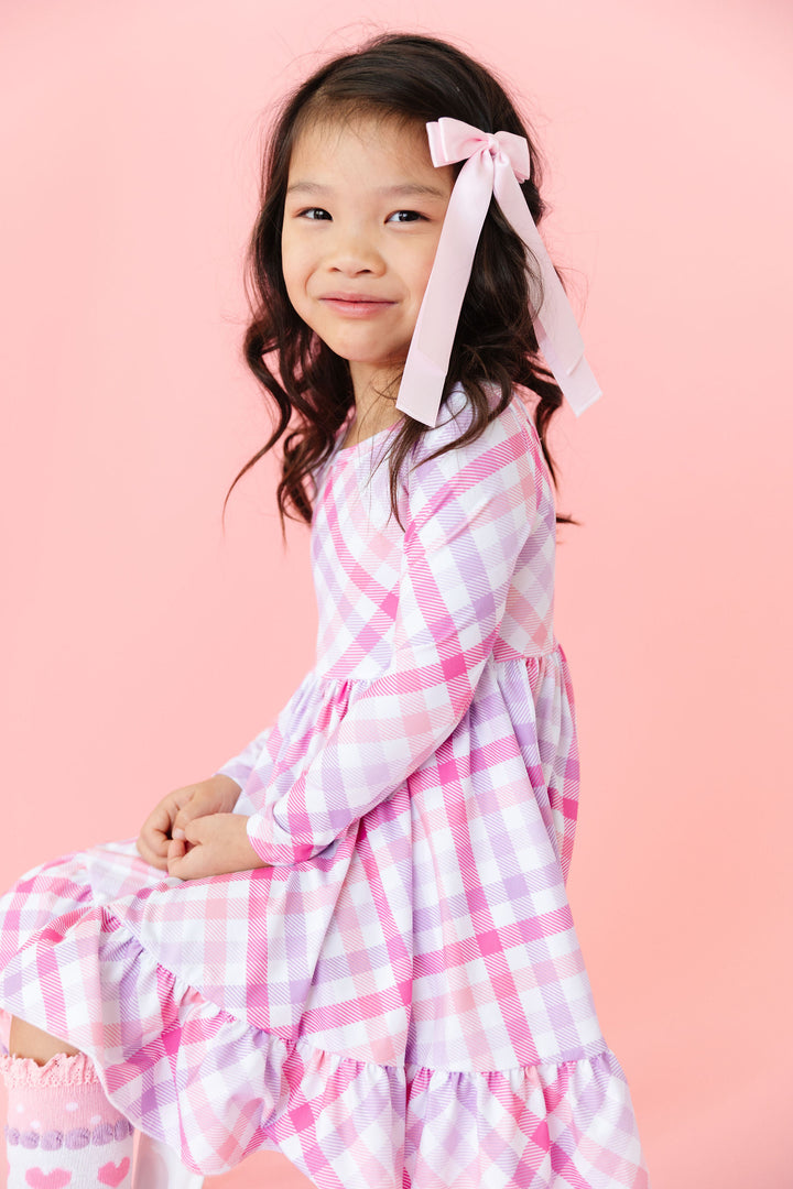 little girl sitting in pink, white and lavender gingham print Valentine's day dress with matching light pink satin bow