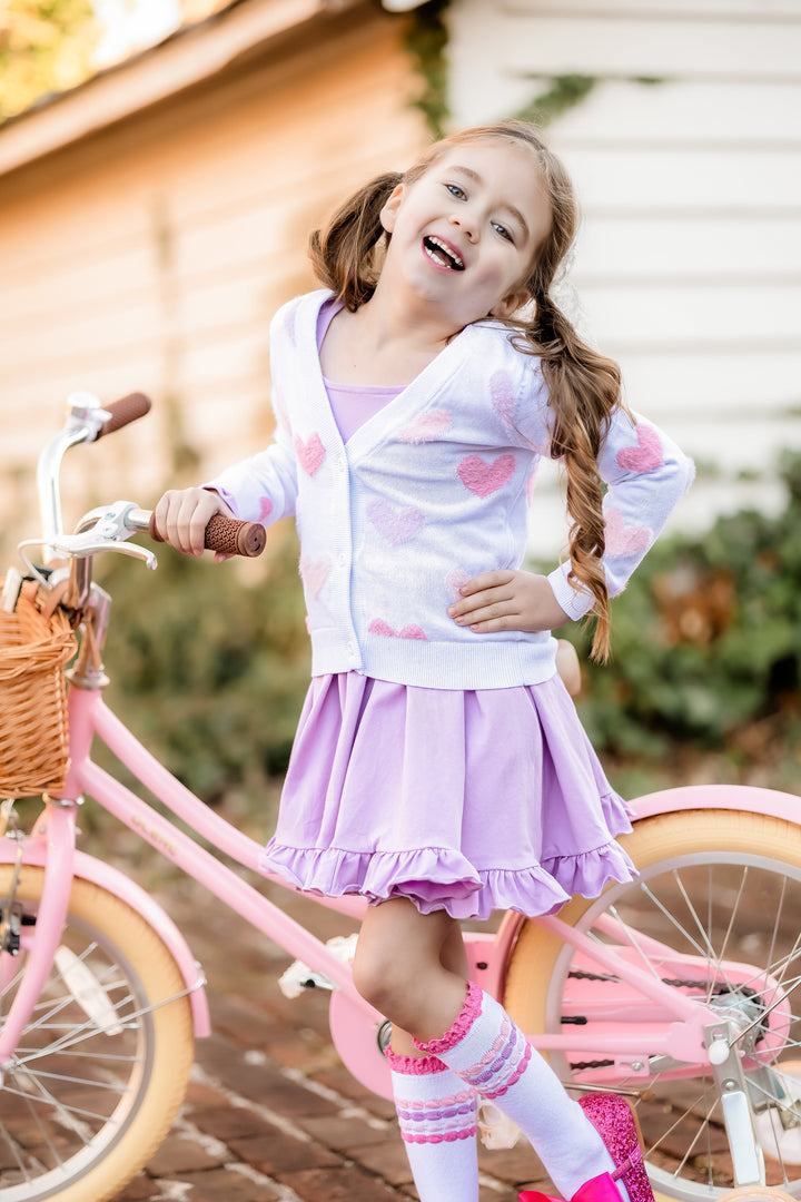 little girl with pink bicycle wearing lavender twirl dress layered with white cardigan with pastel fuzzy hearts and pastel bubble striped knee socks 