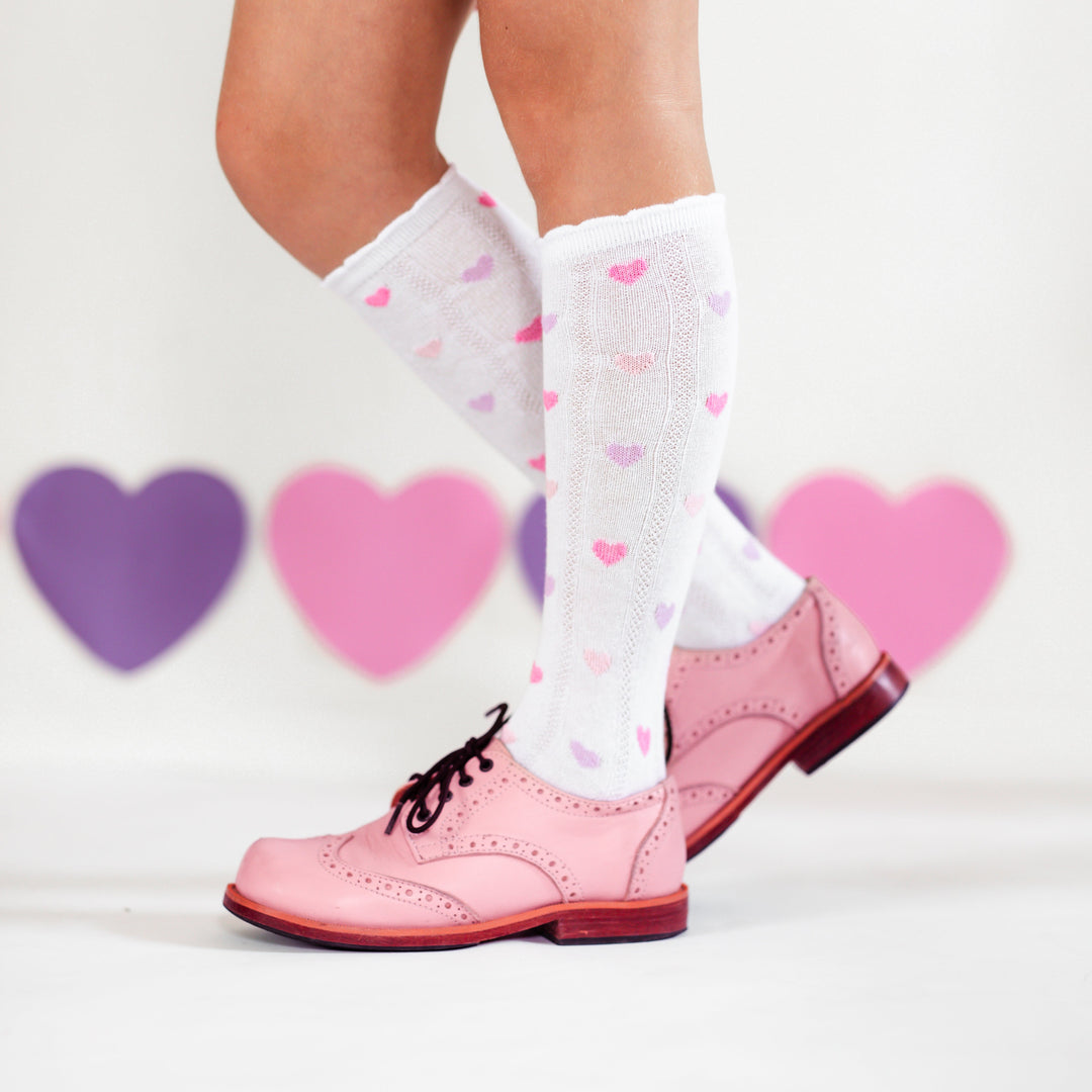 girl in pink saddle shoes in front of heart backdrop wearing white scallop trim knee highs with pastel pink and purple hearts