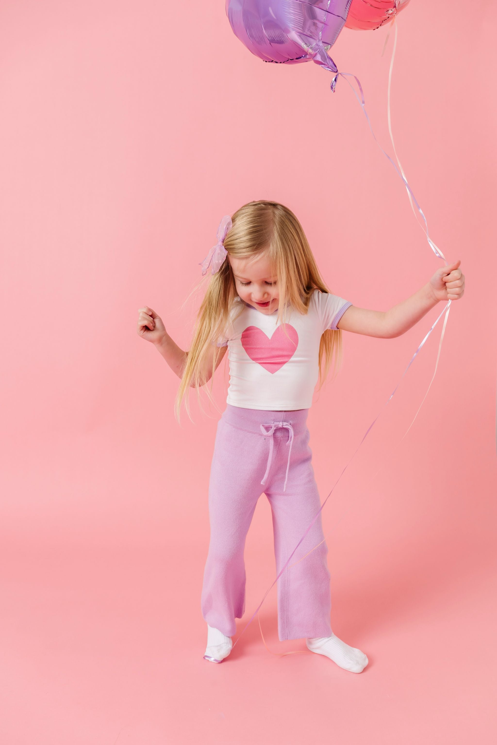 little girl holding balloons in white with pink heart valentines top, lavender knit sweater pants and white socks