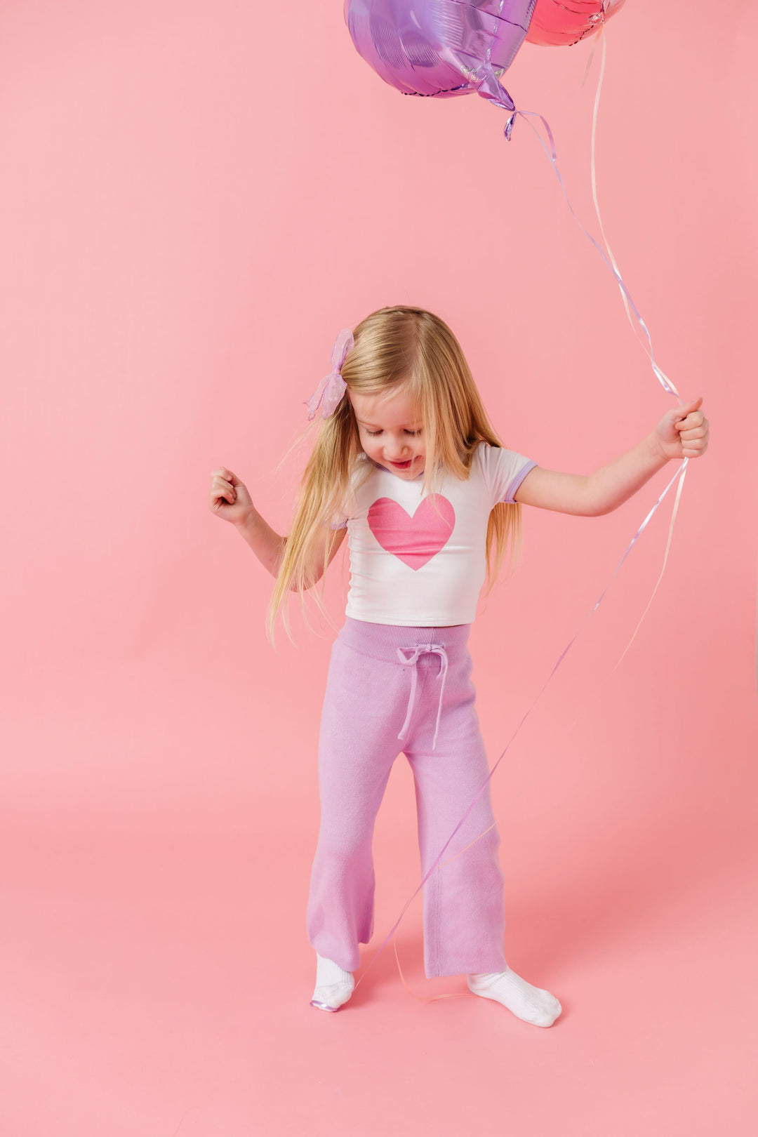 little girl holding balloons in white with pink heart valentines top, lavender knit sweater pants and white socks
