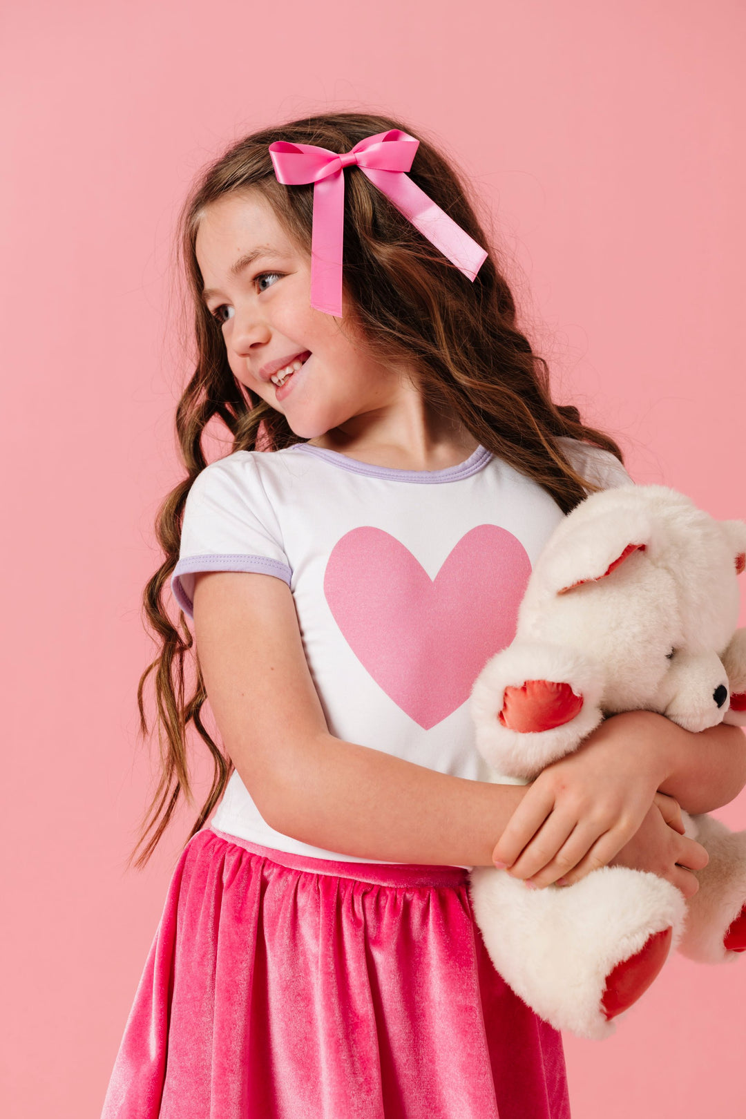young girl holding teddy bear in white bamboo stretchy fitted top with pink heart design and lavender trim paired with pink satin hair bow and matching pink velvet skort