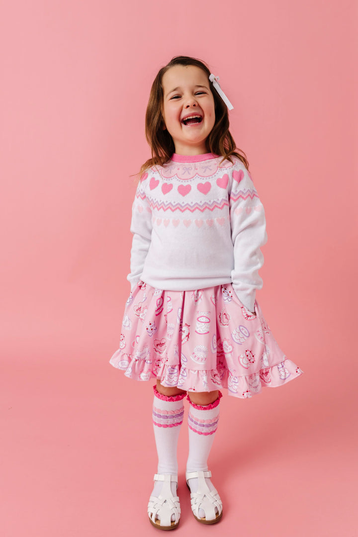 little girl in pink, white and lavender fair isle sweater layered over pink valentines dress featuring heart shaped cakes and matching pastel striped lace top knee highs