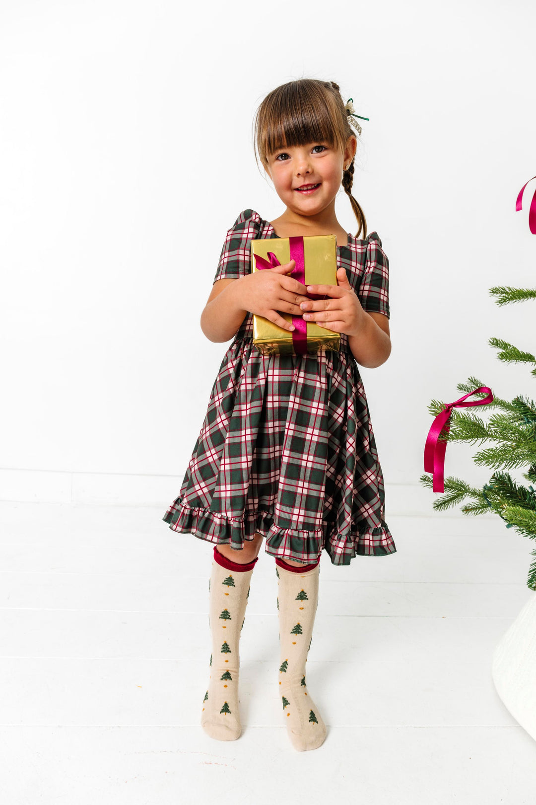 little girl holding Christmas gift in forest green plaid twirl dress with Christmas tree print cream knee highs