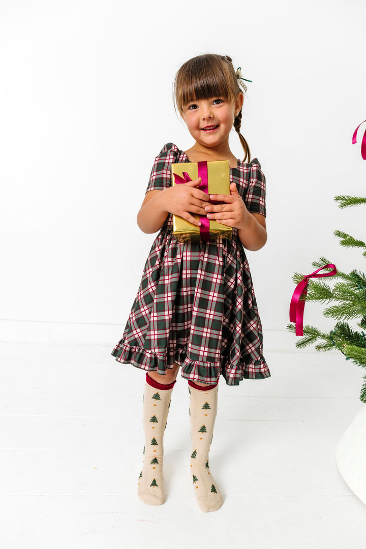 little girl holding Christmas gift in forest green plaid twirl dress with Christmas tree print cream knee highs
