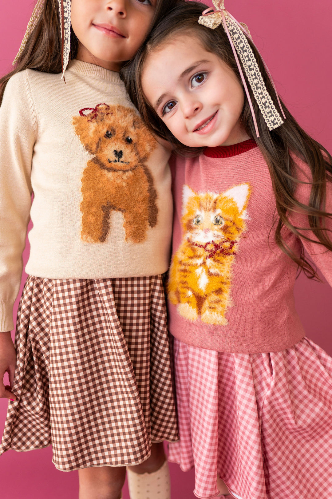 two little girls wearing knit sweaters with matching gingham dresses.  left sweater has doodle dog on it and right sweater has fluffy orange cat graphic