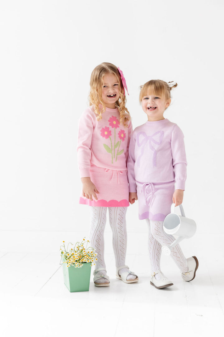 sisters in spring sweater sets with white pointelle tights; one little girl in light pink pullover sweater with floral bouquet design and matching pink skirt with scalloped print trim and another girl in two tone purple bow sweater and matching scallop design skirt