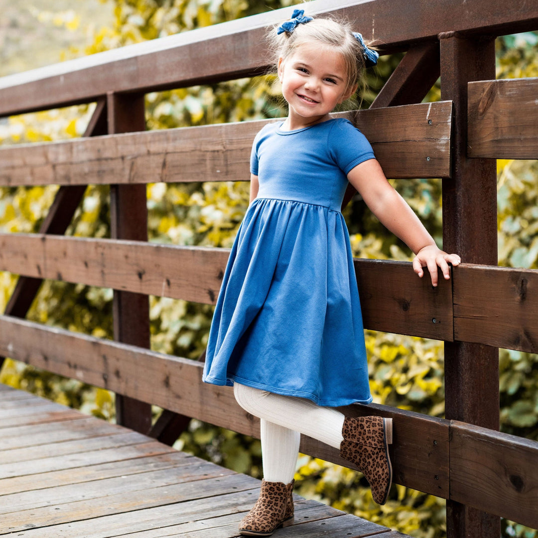 little girl standing against wooden fence wearing denim blue cotton dress and ivory tights