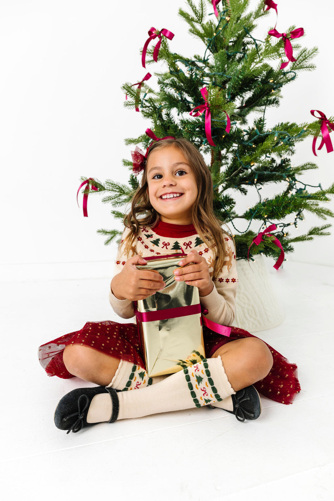 little girl holding gift in front of Christmas tree in cream, crimson and forest green fair isle print sweater and socks with matching tutu bow and skirt