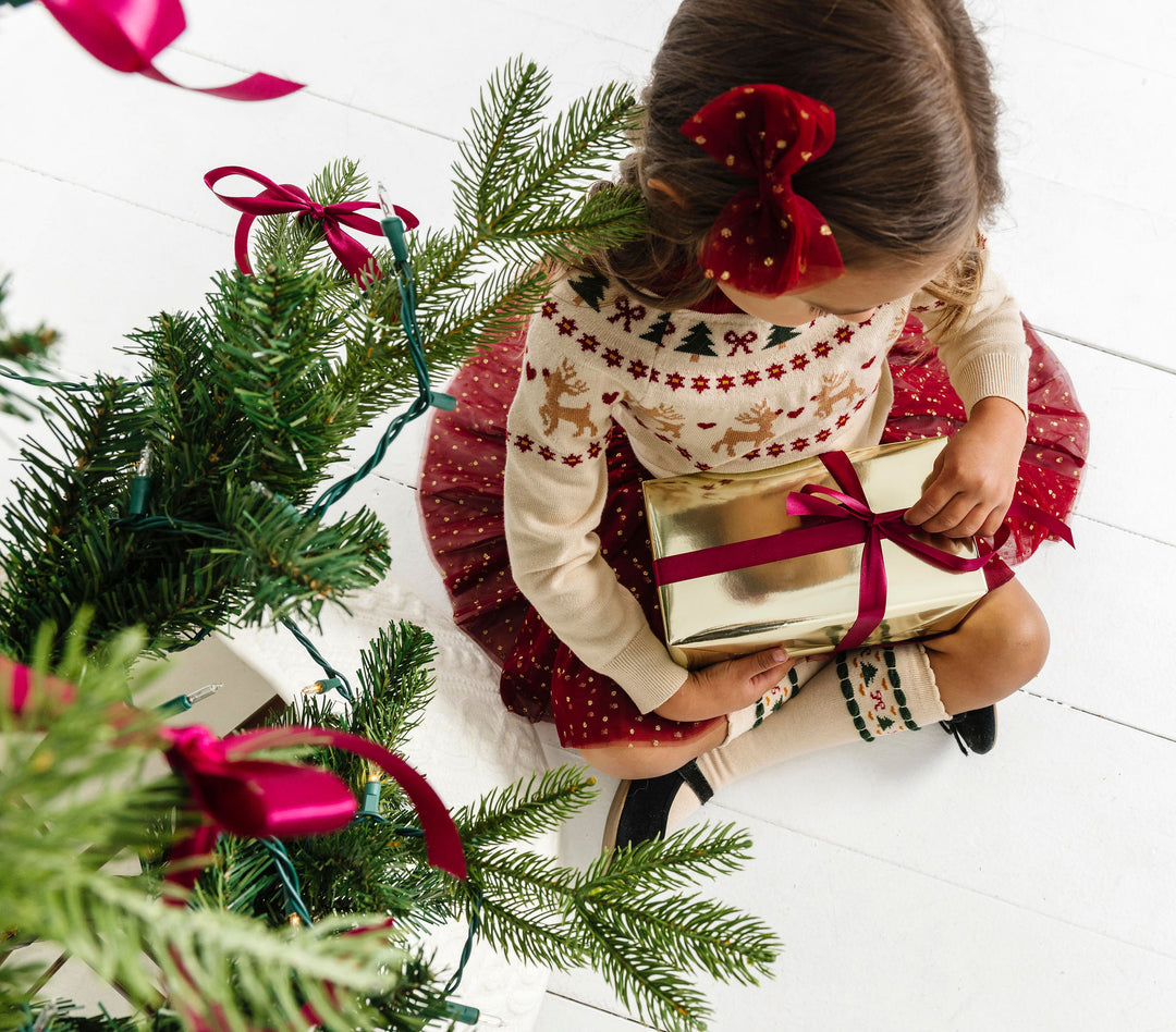 little girl opening gift by Christmas tree in cream fair isle print sweater with coordinating tutu skirt, bow and matching knee-high socks