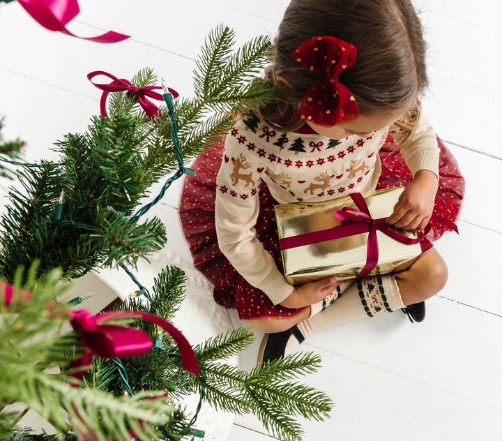 little girl opening gift by Christmas tree in cream fair isle print sweater with coordinating tutu skirt, bow and matching knee-high socks