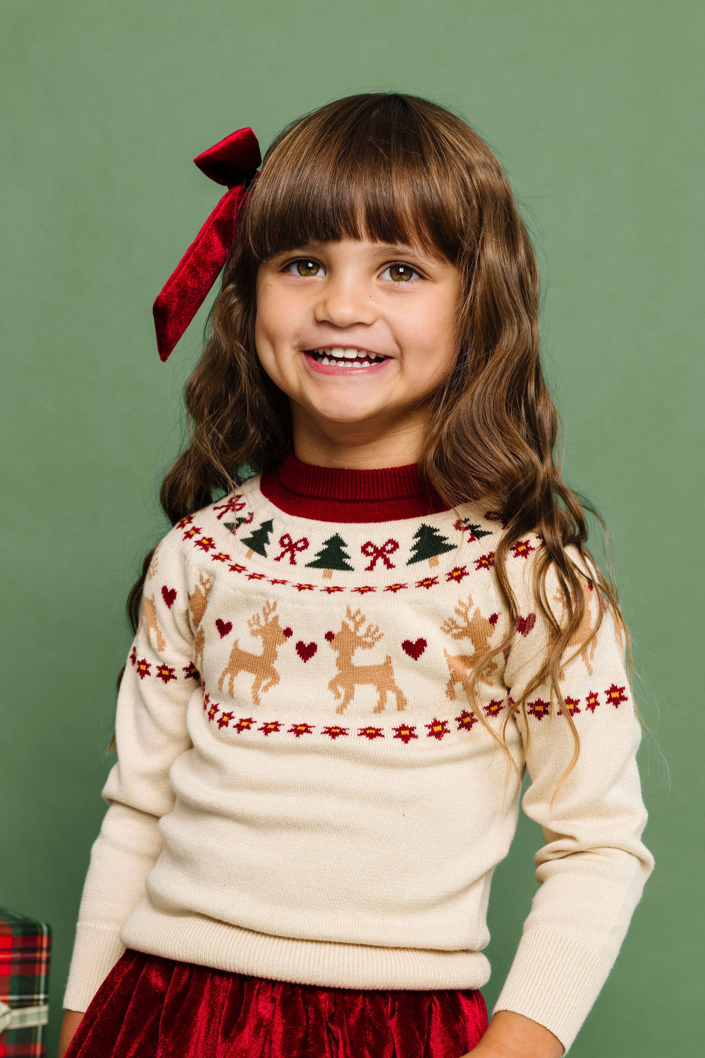 little girl in cream faire isle sweater with reindeer, bow and tree design paired with crimson velvet skirt and matching bow for Christmas