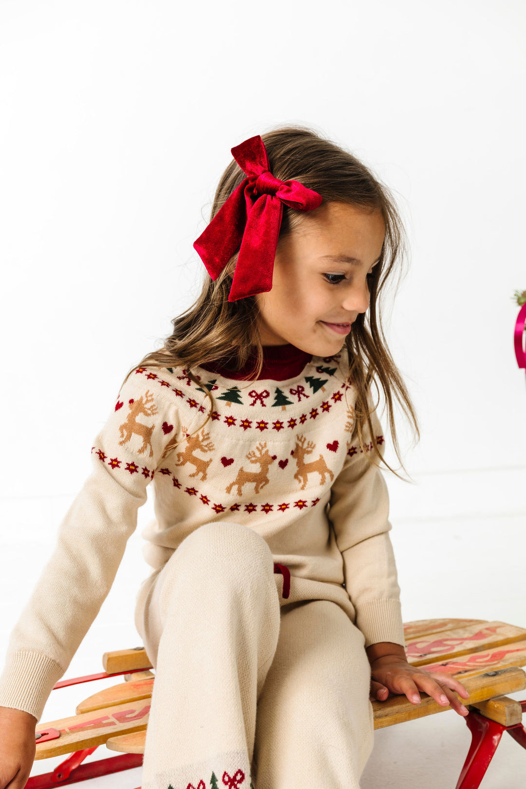 little girl sitting on sled in in cream fair isle sweater and pant set with reindeer design and matching crimson velvet bow on clip for Christmas and winter