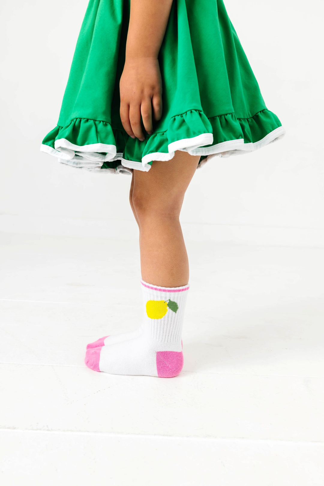 little girl in grass green twirl dress with white trim paired with white crew socks featuring lemon design