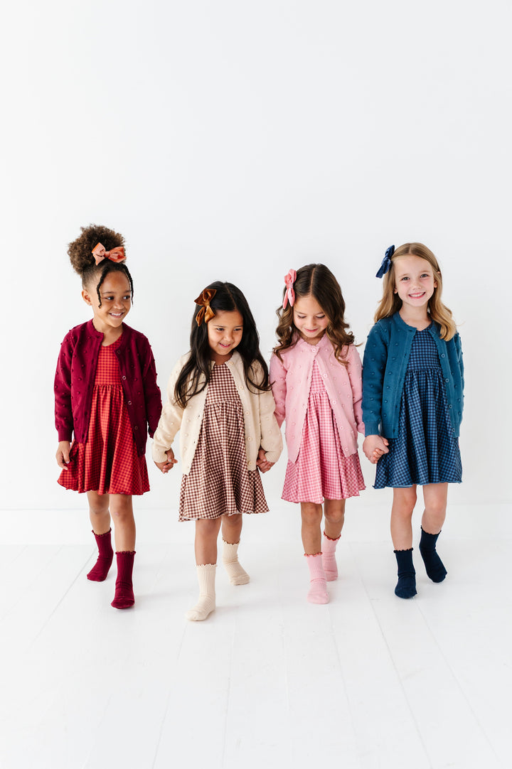 four girls wearing gingham twirl dresses in rust, brownie, old rose and blue paired with matching sweaters, socks and bows