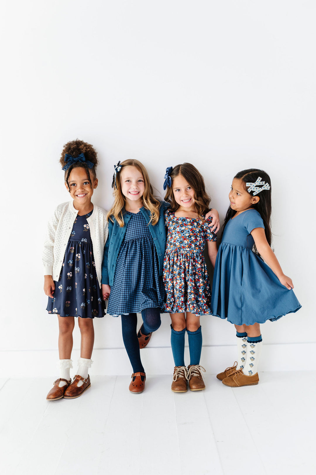 Four young girls standing together against a clean white studio wall, each wearing a different blue-toned dress or outfit. The outfits include navy, blue, floral multicolor, and solid medium blue, with brown shoes and white socks.