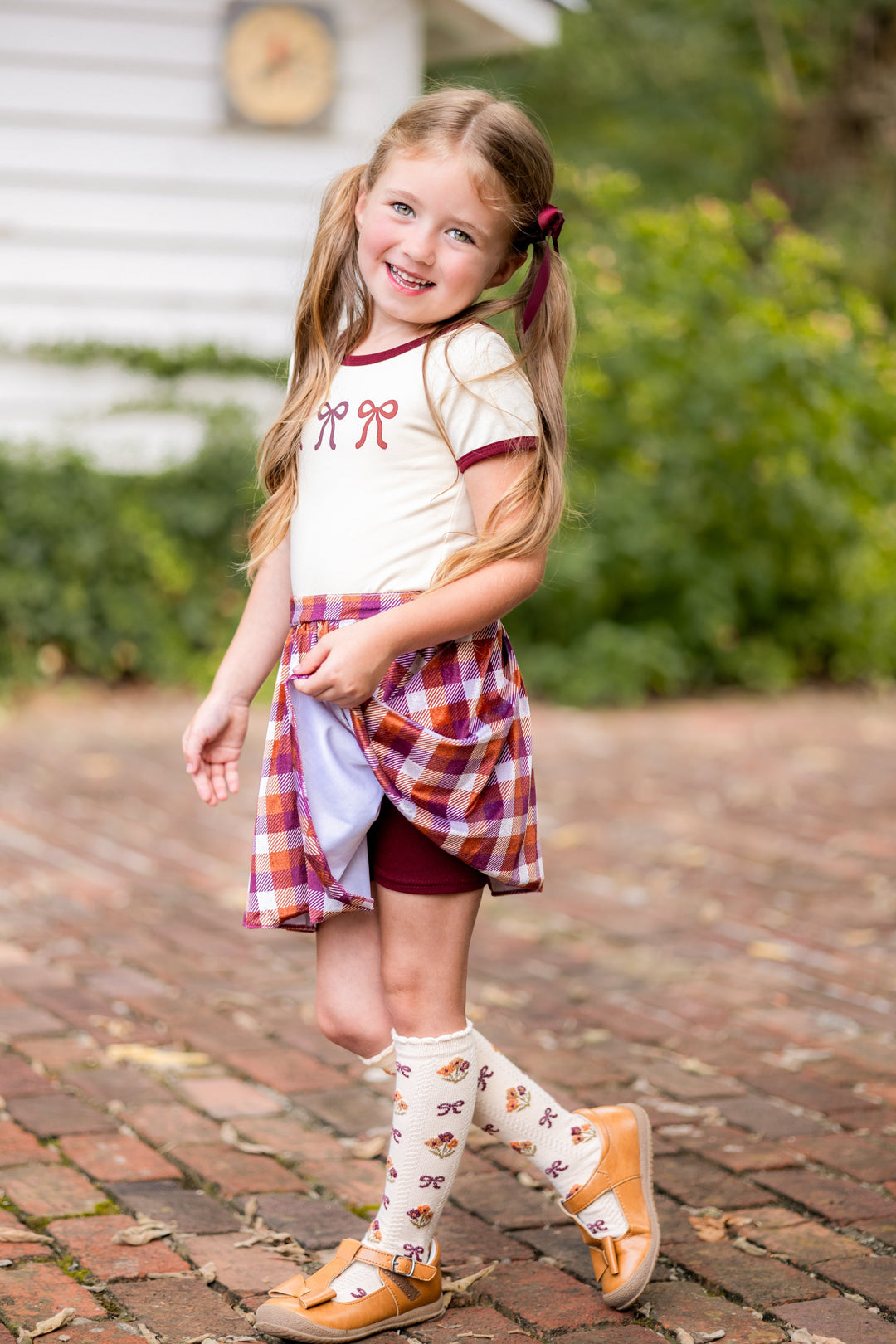 little girl in cream fitted top with bow print, matching fall plaid velvet skort with built in burgundy shorts, cream floral knee highs and burgundy satin bow