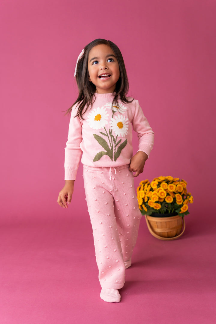 little girl walking on on pink background wearing light pink daisy design sweater paired with matching pink knit bubble textured pants
