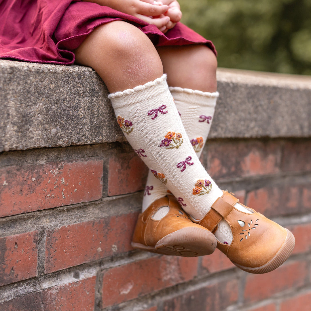 little girl sitting on brick wall in cream knee high socks with fall bow pattern and bows paired with matching burgundy cotton blend skort