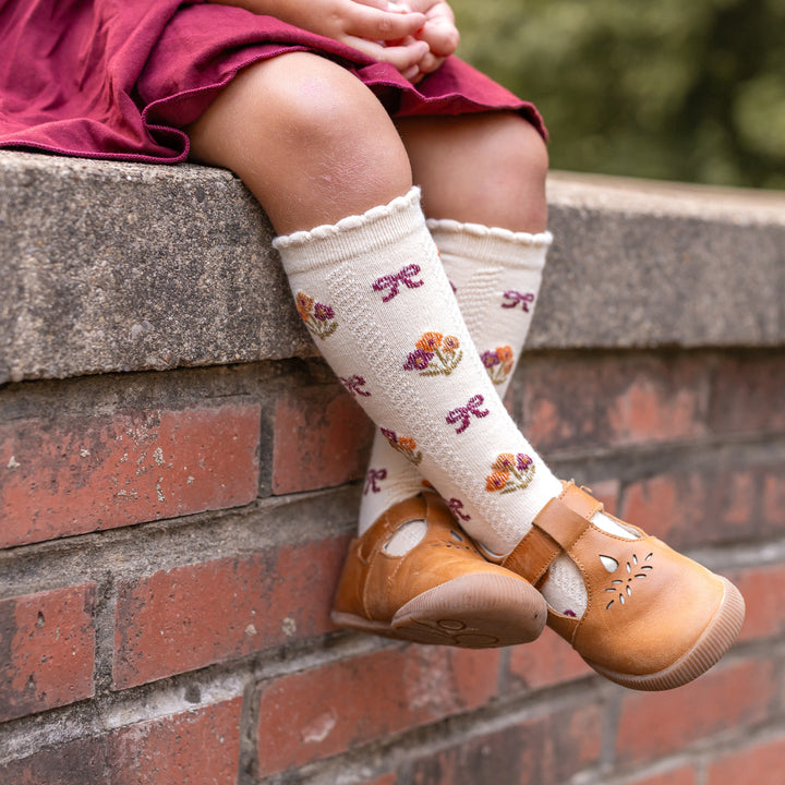 little girl sitting on brick wall in cream knee high socks with fall bow pattern and bows paired with matching burgundy cotton blend skort