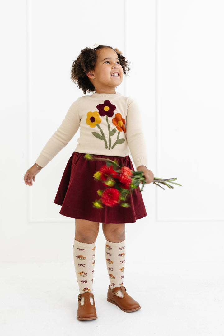 little girl holding fall flowers in cream pullover knit flower sweater, burgundy cotton blend skort and cream scalloped knee highs with matching flower design 